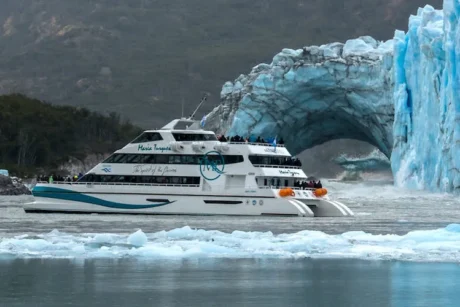 Una embarcación turística de dos pisos, blanca y azul, navega cerca de un glaciar con un arco de hielo visible. La escena muestra el barco, con pasajeros en cubierta, rodeado de agua grisácea y témpanos de hielo flotantes, con una montaña cubierta de vegetación al fondo.