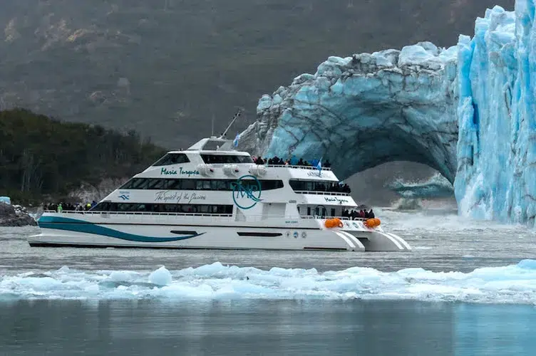 Una embarcación turística de dos pisos, blanca y azul, navega cerca de un glaciar con un arco de hielo visible. La escena muestra el barco, con pasajeros en cubierta, rodeado de agua grisácea y témpanos de hielo flotantes, con una montaña cubierta de vegetación al fondo.