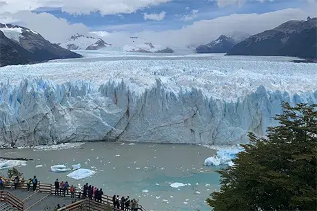 Gran imagen panorámica del glaciar Perito Moreno en Argentina, con su imponente frente de hielo azul y blanco que se eleva sobre el lago. Al pie del glaciar, en las pasarelas de madera, se observa a un grupo de turistas maravillados por el espectáculo natural. El paisaje se completa con montañas nevadas y un cielo nublado que resalta la majestuosidad de este atractivo turístico de la Patagonia.